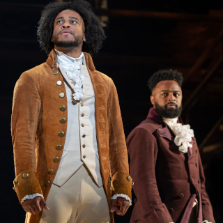 Two men in historical costumes stand on stage. One wears a tan coat and beige vest with ruffled shirt, looking upward; the other, in a dark maroon coat, looks forward. Both appear serious, as if in a dramatic performance.