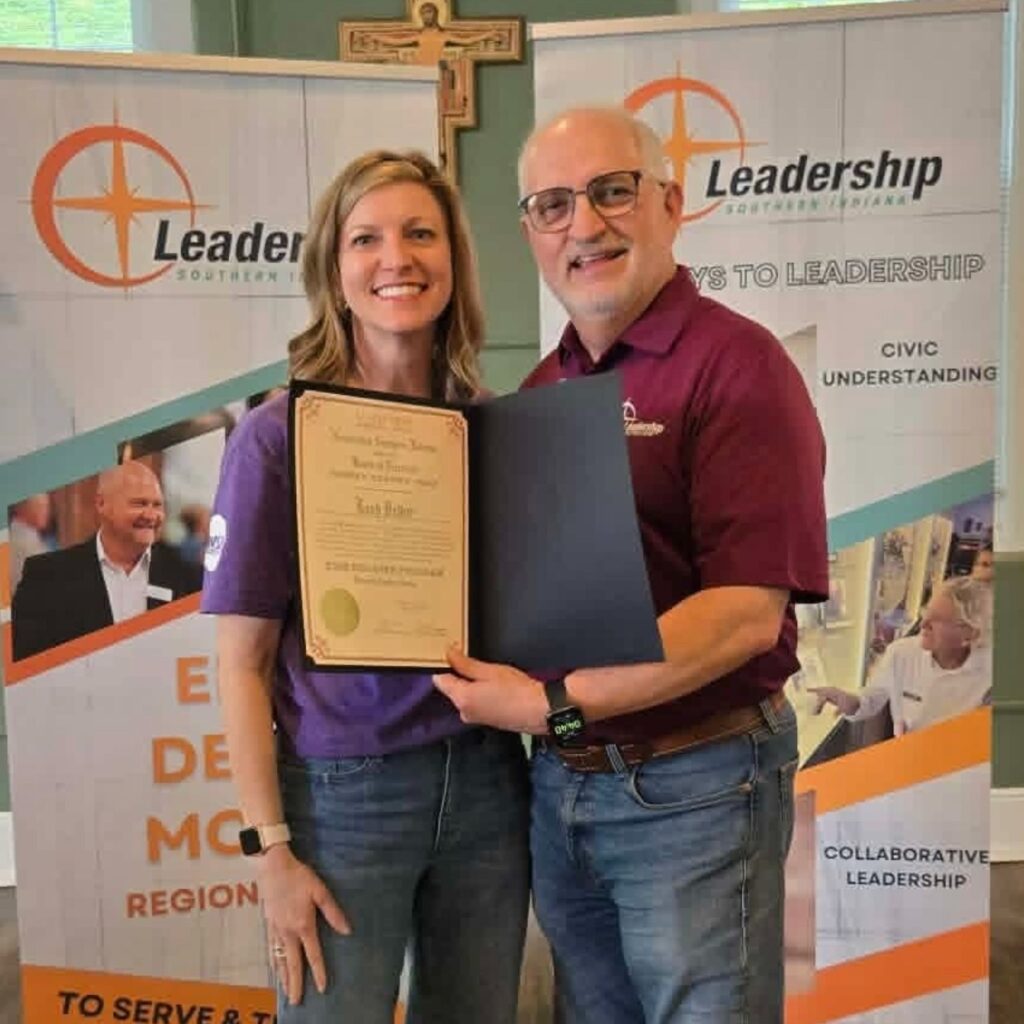 Two people smile while holding a certificate in front of Leadership Southern Indiana banners. A cross is mounted on the wall behind them, and both are wearing casual clothes.