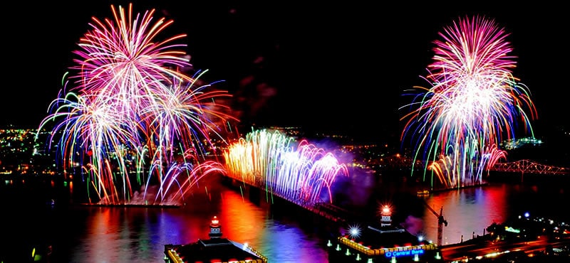 Colorful fireworks explode over a cityscape at night, illuminating a river and a large bridge below. Bright reflections shimmer on the water, and city lights glow in the background.