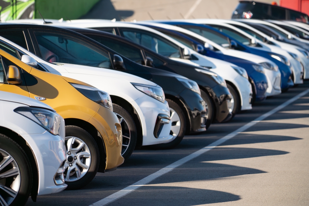 A row of parked cars in various colors, including yellow, white, black, and blue, lined up diagonally in a parking lot on a sunny day.