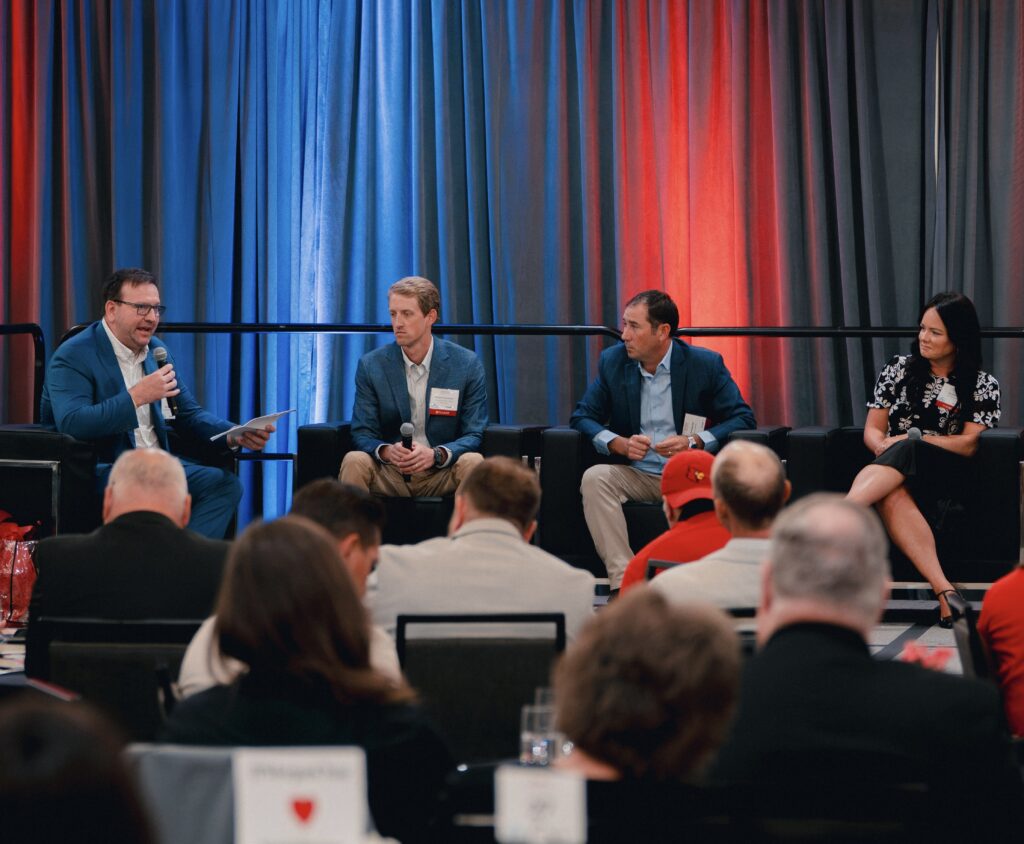 Four people sit on a stage, engaging in a panel discussion in front of an audience. The background features red and blue curtains. Three men and one woman are seated, with one man holding a microphone.