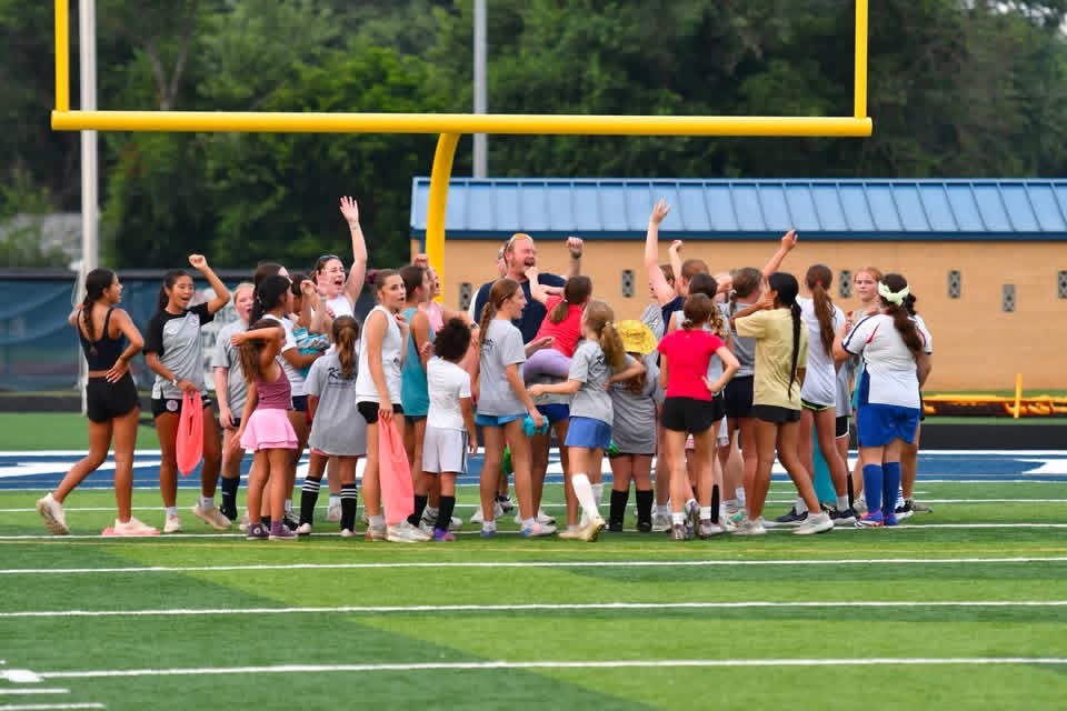 A group of girls and coaches stand together on a football field under a goalpost, some raising their hands, suggesting a team huddle or celebration. Trees and a building are visible in the background.