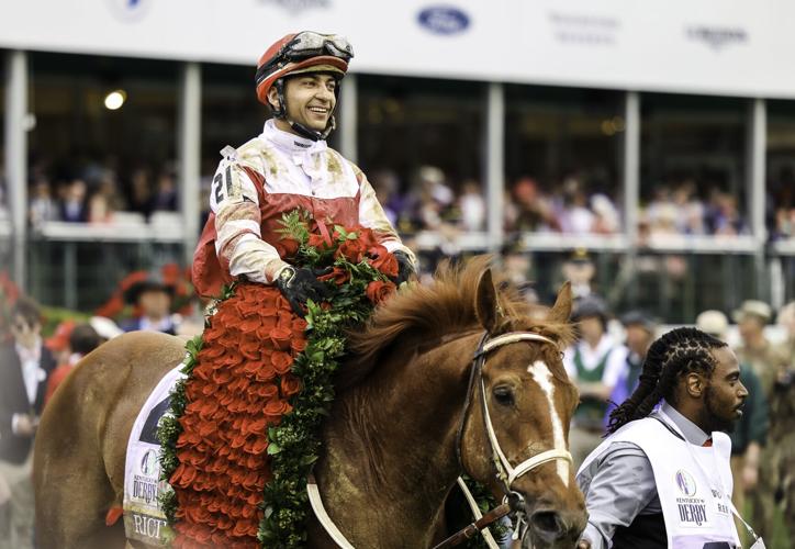 A smiling jockey wearing red and white silk sits on a chestnut horse draped in a garland of red roses, surrounded by people at a crowded racetrack event.