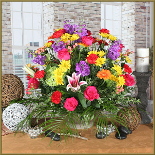 A vibrant floral arrangement with pink roses, purple and yellow daisies, orange gerbera daisies, and greenery sits on a brown table in front of tall windows and brick walls, surrounded by decorative objects.