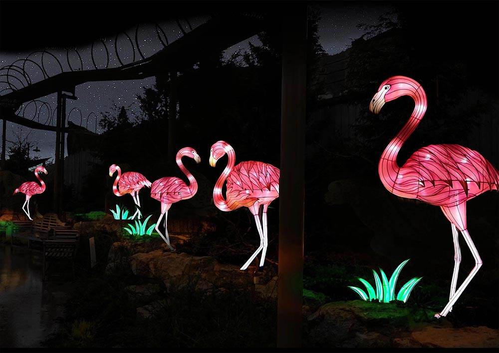Four illuminated flamingo sculptures glow pink at night in a garden with rocks and green-lit plants, under a dark, starry sky. Silhouetted trees and wire arches are visible in the background.