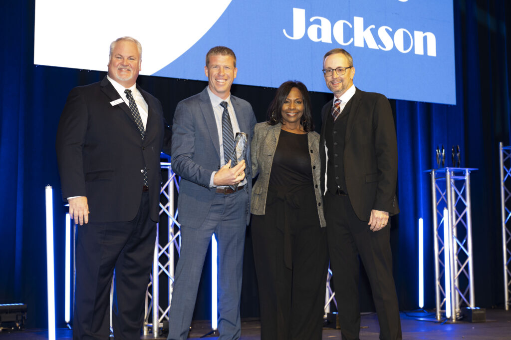 Four people in business attire stand on stage smiling. One person holds a glass award. Behind them is a blue screen with the name Jackson partially visible. Stage lights and metal truss structures are in the background.