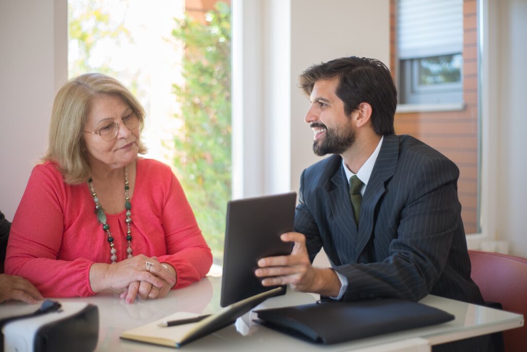 A man in a suit shows a tablet to an older woman in a pink top. They are sitting at a table, engaged in a friendly conversation in a bright, modern office.