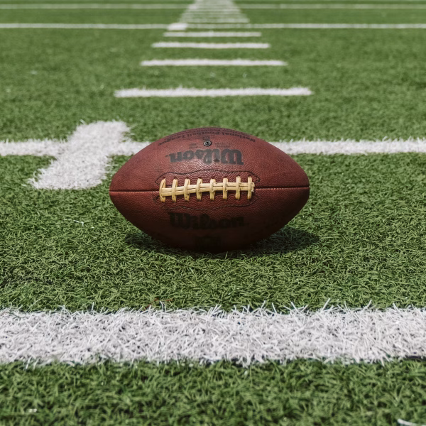 A brown football sits on a green field with white yard lines, positioned near the center of a horizontal white line. The field’s synthetic grass and markings are clearly visible.