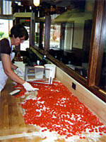 A person wearing an apron and gloves prepares a large batch of red candy pieces on a wooden countertop in a kitchen or candy shop.