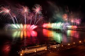 Colorful fireworks light up the night sky over a river, reflecting brightly on the water. A bridge and a docked riverboat are visible in the foreground, with city lights lining the waterfront.