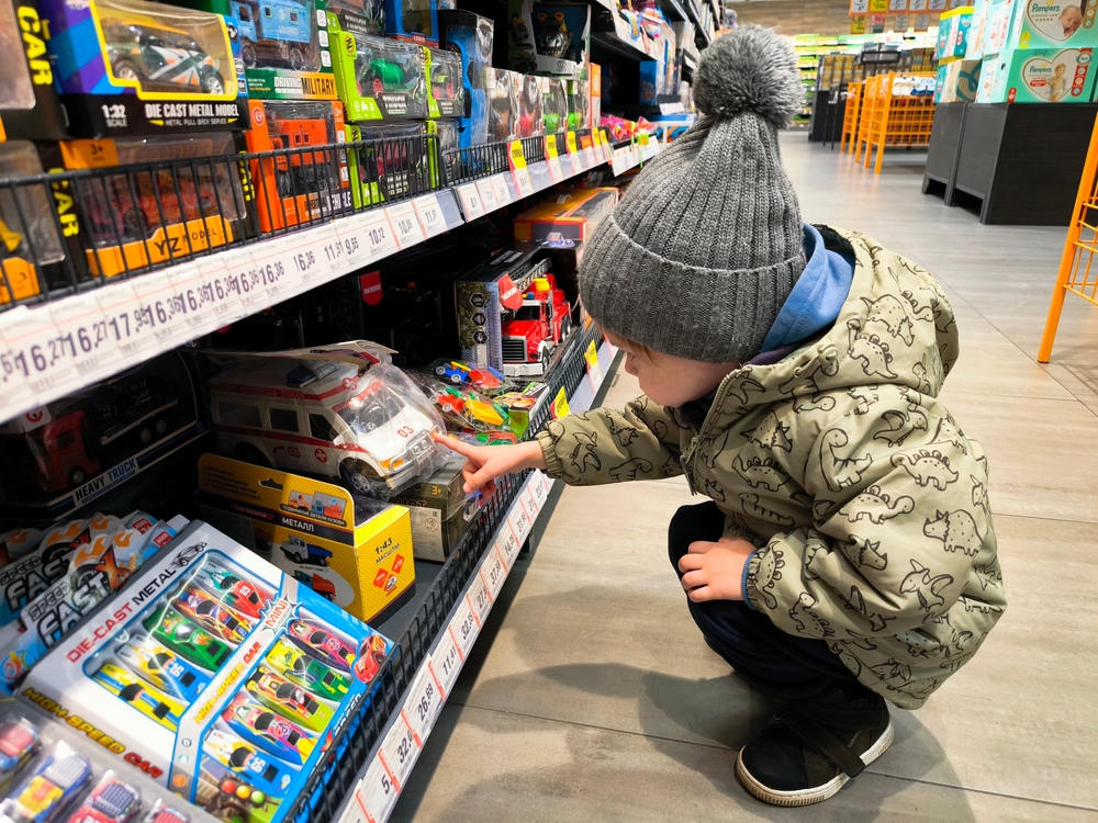 A young child in a gray hat and dinosaur-patterned jacket crouches in a store aisle, reaching for a toy car on a shelf filled with colorful toy vehicles.
