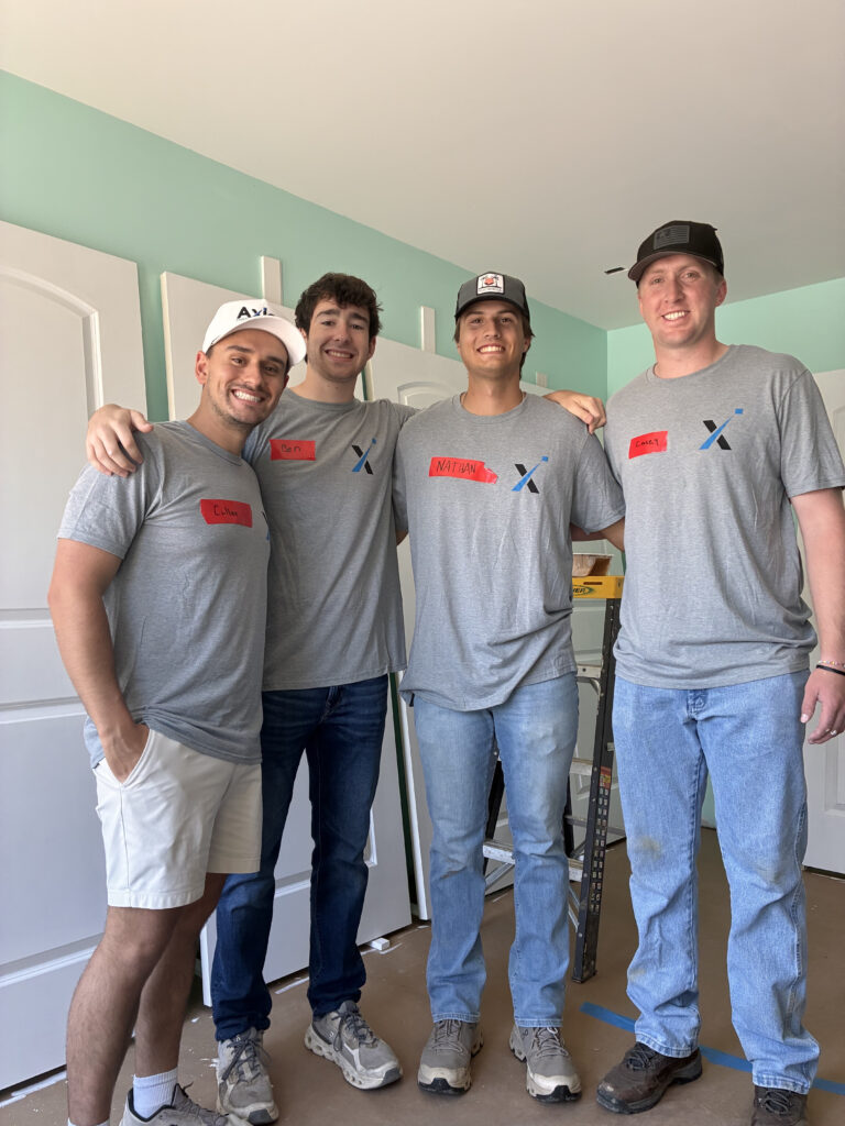 Four smiling men stand indoors, wearing matching gray T-shirts and name tags. They have their arms around each other, and the room appears to be under renovation with construction materials visible.