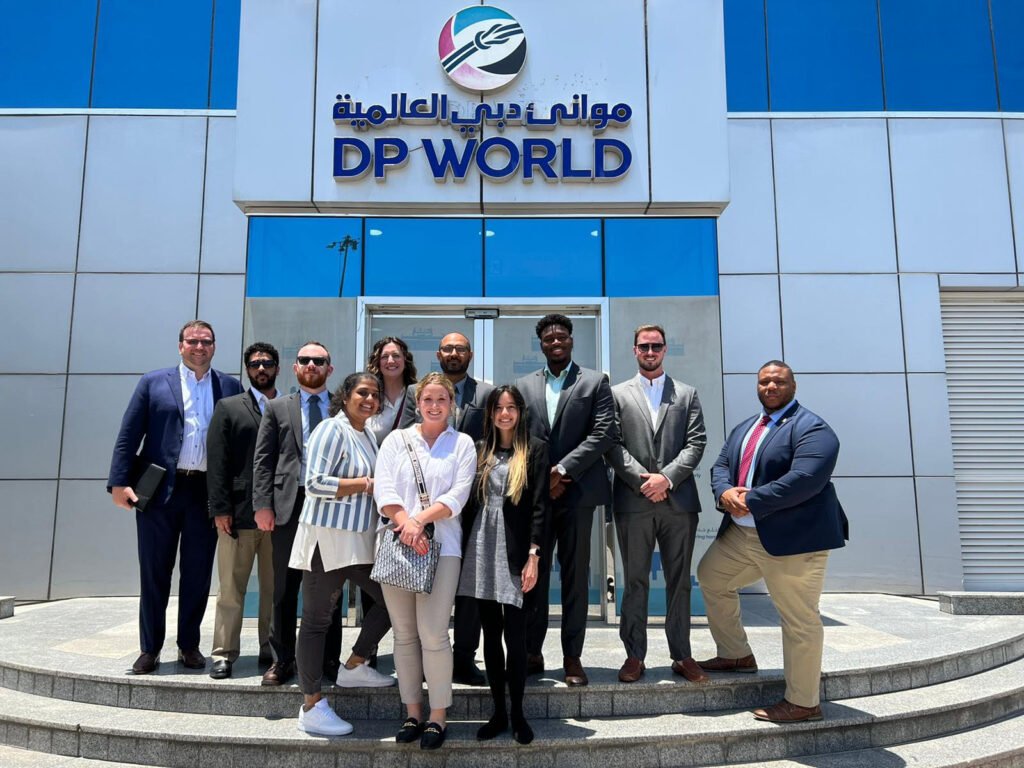 A group of ten people, dressed in business attire, stand smiling in front of the DP World building entrance with Arabic and English signage above them. The building exterior features glass and metal panels.