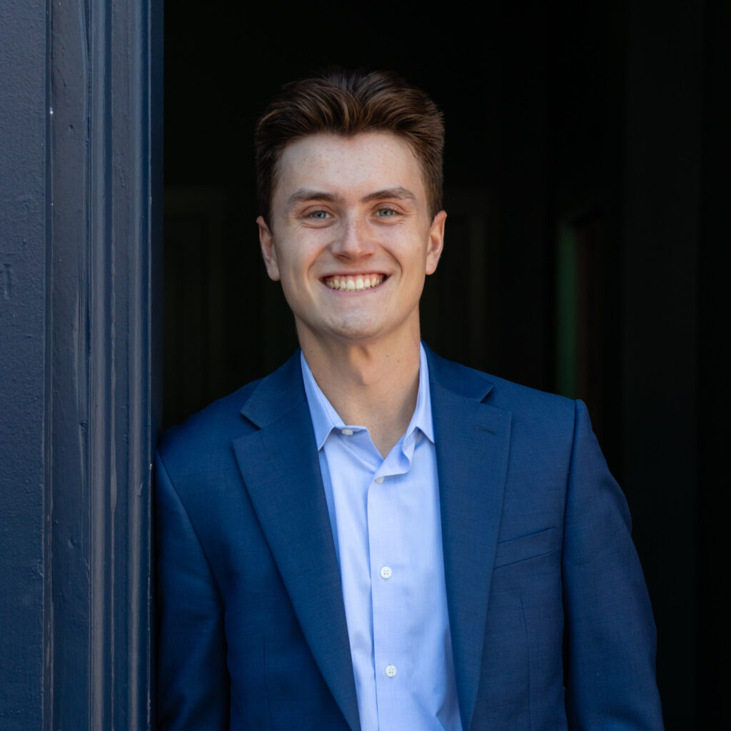 A young man wearing a blue suit and light blue shirt stands in a doorway, smiling warmly at the camera.