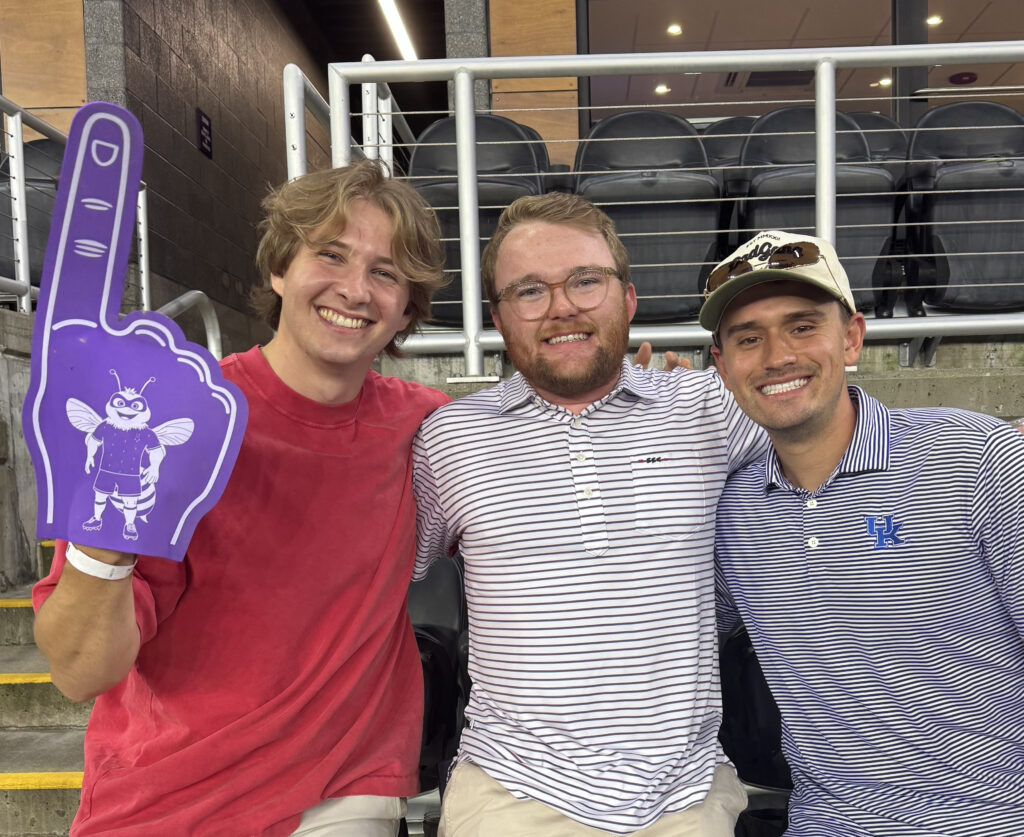 Three smiling young men sit together in stadium seats; one wears a red shirt and holds a purple foam finger with a bee graphic, the others wear striped shirts and caps. Empty seats and metal railings are visible behind them.