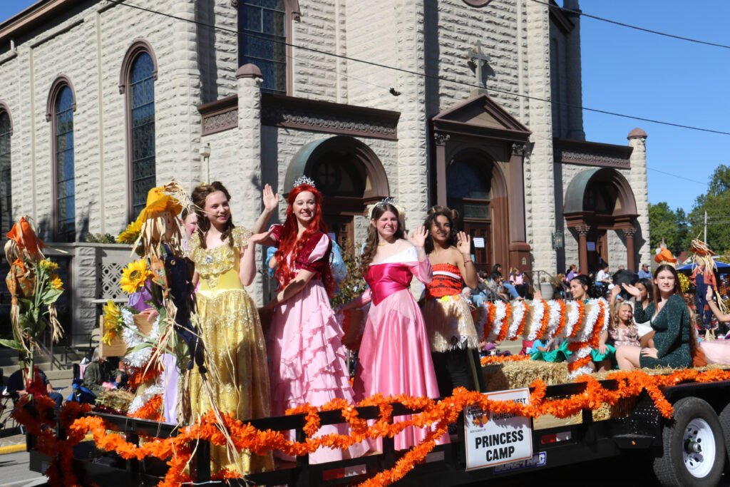 Four women dressed as princesses wave from a parade float decorated with autumn leaves, flowers, and orange garlands in front of a large stone building. A sign on the float reads “PRINCESS CAMP.” People watch from the sidewalk.