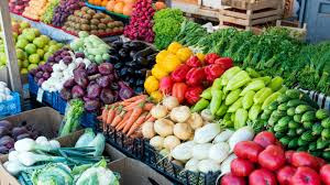 A colorful display of assorted fresh vegetables, including carrots, peppers, tomatoes, eggplants, and leafy greens, arranged in rows at an outdoor market stall.