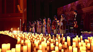 A musical ensemble performs on stage surrounded by numerous glowing candles, creating a warm and intimate atmosphere. The musicians are dressed formally and appear to be playing string instruments.