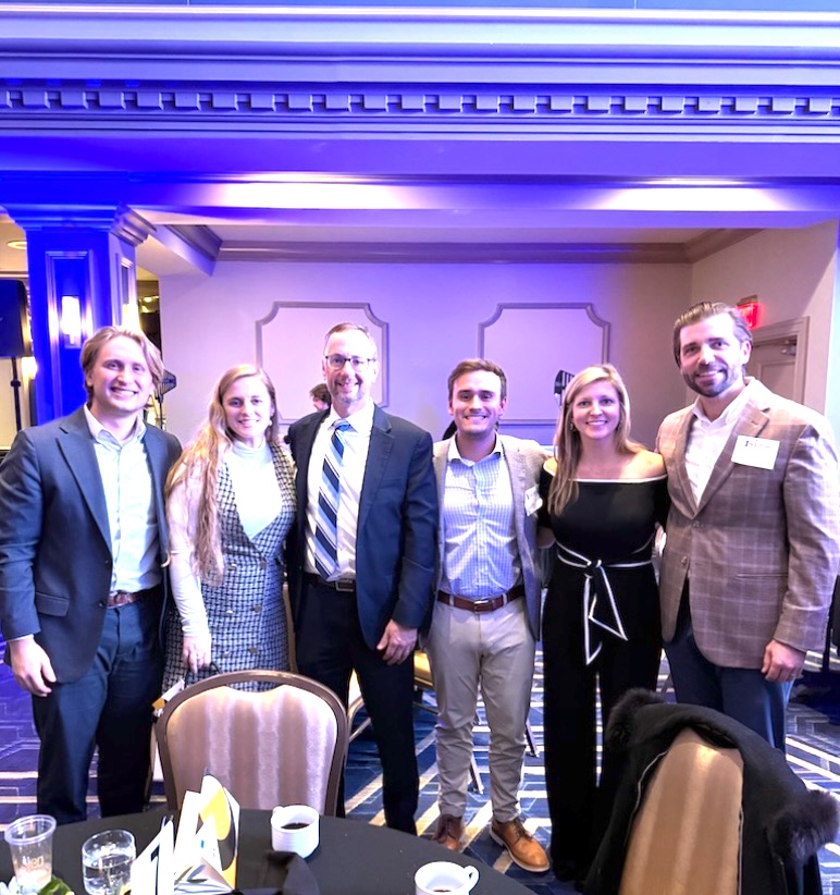 Six people dressed in business attire stand closely together, smiling for a group photo at an indoor event or conference. They are posed in front of a table and under blue lighting in a formal room.